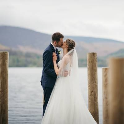The couple kiss on a wooden jetty with a lake and hills behind them.