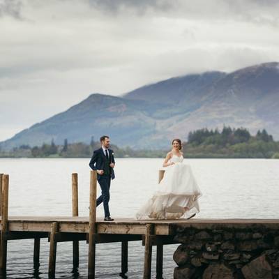 The couple walk along a jetty on the lake with a cloud-topped mountain behind.