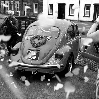 Confetti in the air around a vintage Beetle with a just married sign while a man takes a photo, black and white.