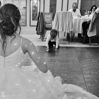 A young boy crouches on the dance floor with a toy while the bride's dress fills the foreground, black and white.