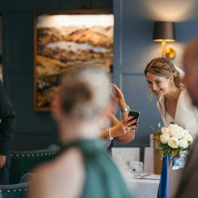 The bride laughs with her bouquet while a guest takes a photo on a phone.